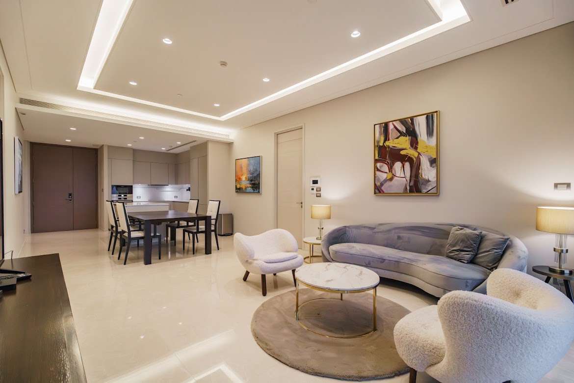 Open-plan modern living and dining area with a curved gray sofa, white armchairs, and a marble coffee table in a well-lit room.